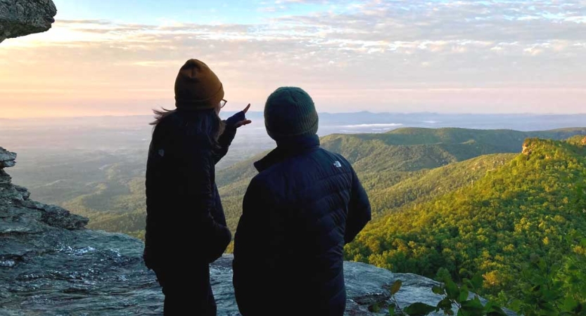 Two people with their backs to the camera stand on a rocky overlook, viewing the vast blue ridge mountains below. 